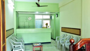 A reception area with a green counter, white plastic chairs, a wooden table with magazines, and a ceiling fan. - Sithara Guest House (Palakkad)