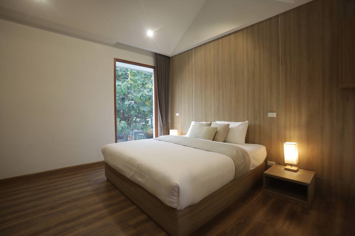 A modern bedroom with a large bed, wooden flooring, a nightstand with a lamp, and a window with a view of greenery.