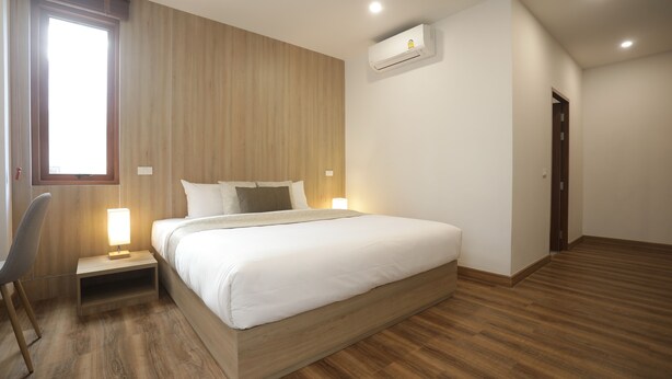 A modern hotel room with a wooden floor, a bed with white linens, a nightstand with a lamp, and an air conditioning unit on the wall. - Kyoto Great Hotel (Chiang Mai)