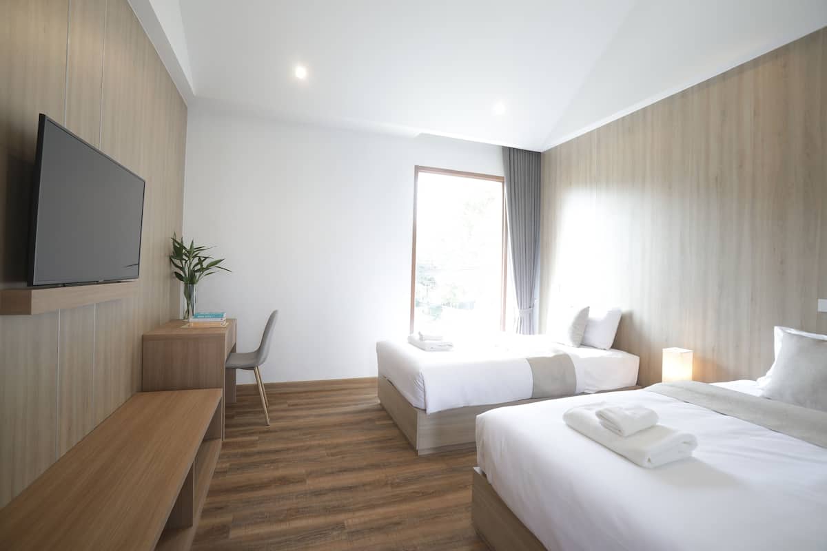 A modern hotel room with a wooden floor, a flat-screen TV mounted on the wall, a desk with a chair, two beds with white linens, and a large window with curtains.
