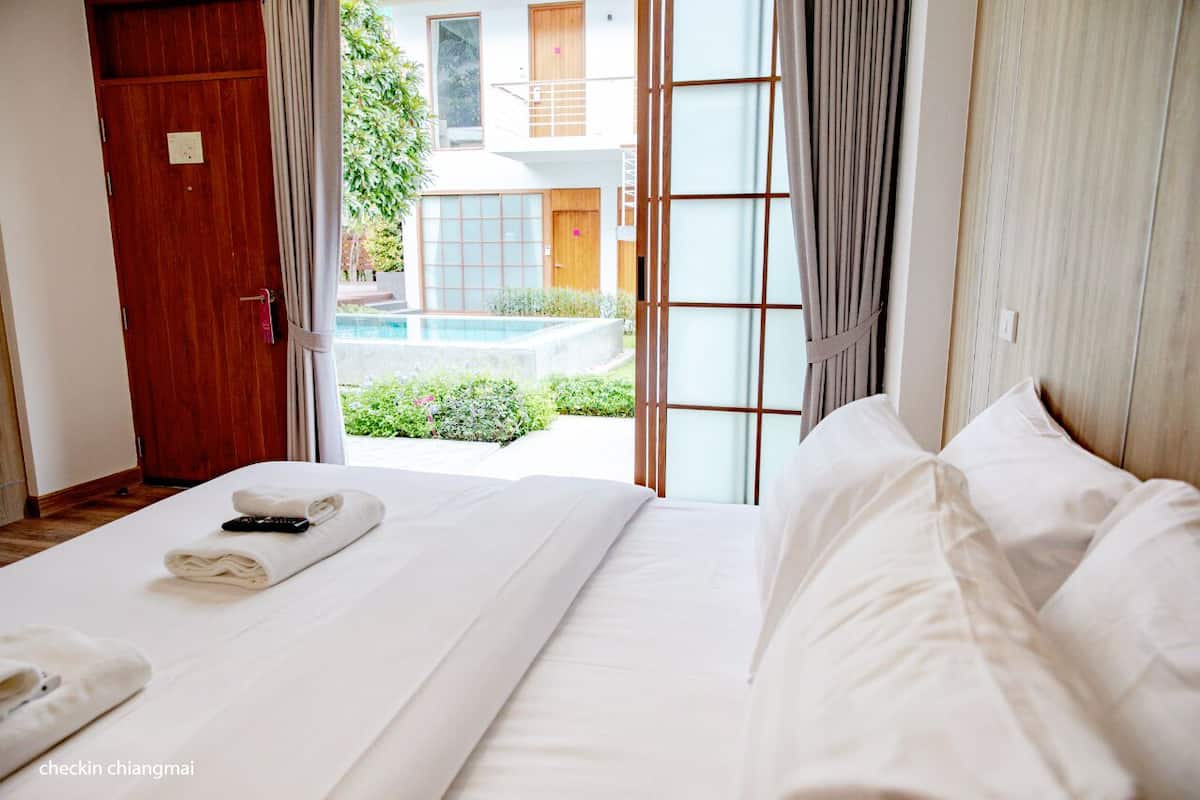 A hotel room with a large bed, a wooden door, and a view of a swimming pool and greenery outside.