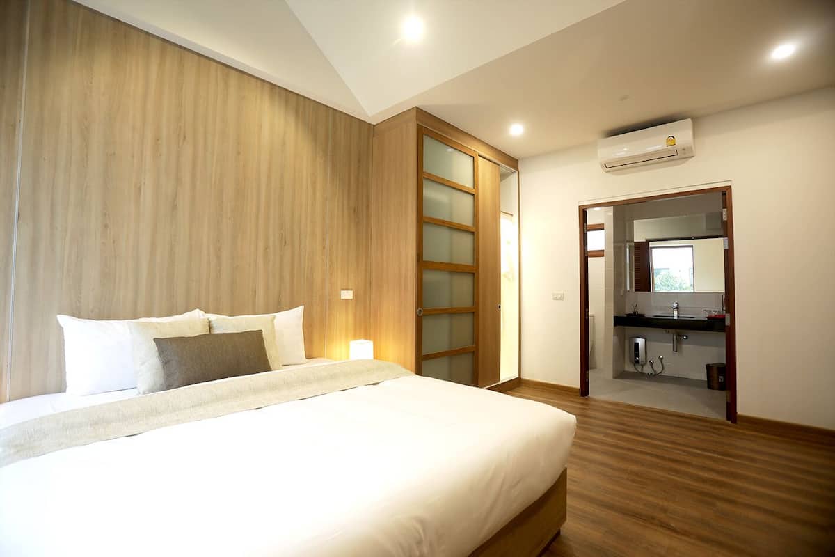 A modern bedroom with a large bed, wooden panel walls, a sliding glass door, and a bathroom visible through an open door.