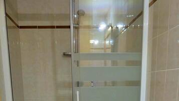 Une salle de bain avec une douche en verre, des toilettes dont l’abattant est endommagé et des murs carrelés.