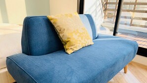 A blue sofa with a yellow pillow in a room with wooden flooring and a window. - WELLSTAY　Osaka　Honmachi　Apartment hotel (Osaka)