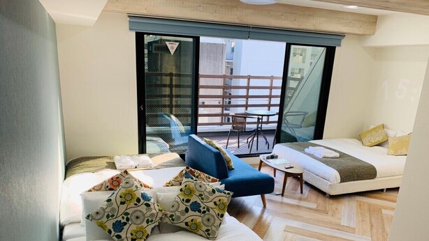 A hotel room with a bed, a sofa, a chair, and a small table. There is a balcony with a railing and a view of the outside. - WELLSTAY　Osaka　Honmachi　Apartment hotel (Osaka)