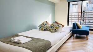 A neatly made bed with a green blanket and white pillows, a blue chair, and a view of buildings through a window. - WELLSTAY　Osaka　Honmachi　Apartment hotel (Osaka)