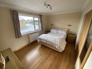 A bedroom with a large bed, a window with curtains, a radiator, a wooden floor, and a nightstand with a lamp.