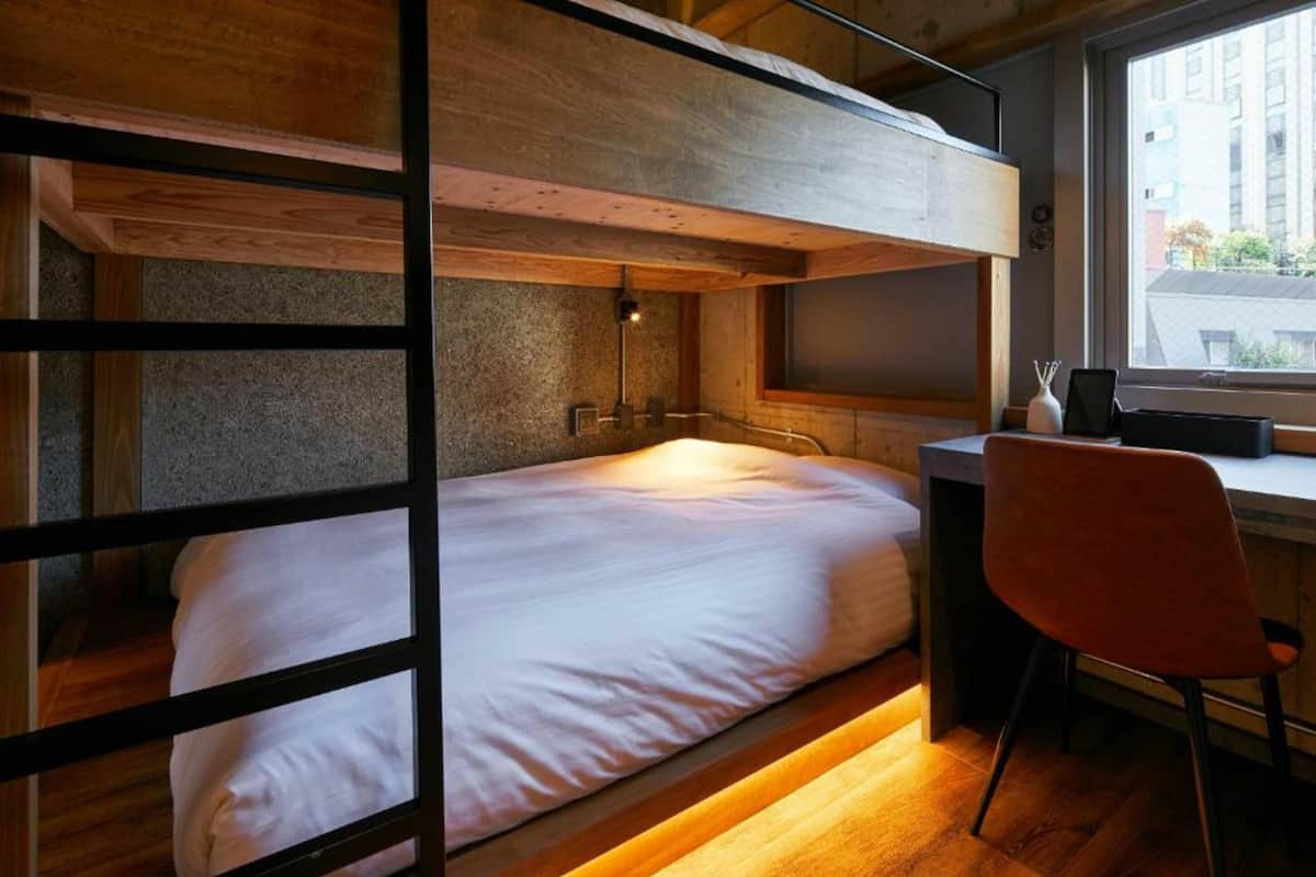 Ein Etagenbett mit Schreibtisch und Stuhl, ein Fenster mit Blick auf Gebäude und eine Lampe auf dem Schreibtisch.