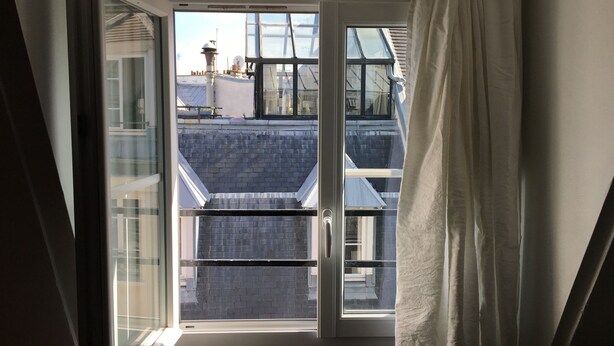 Een kamer met een groot raam, een gordijn en uitzicht op gebouwen.