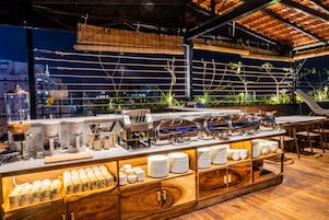 Buffet montado com diversos pontos de comida, uma máquina de café e um balcão de madeira com pratos empilhados.