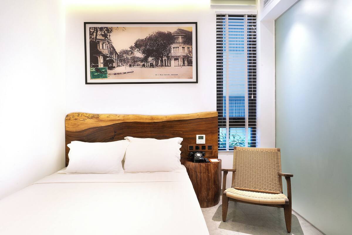 Una cama bien hecha con sábanas blancas, un cabecero de madera, un cuadro en la pared, una silla de mimbre y una mesita de madera con un teléfono.