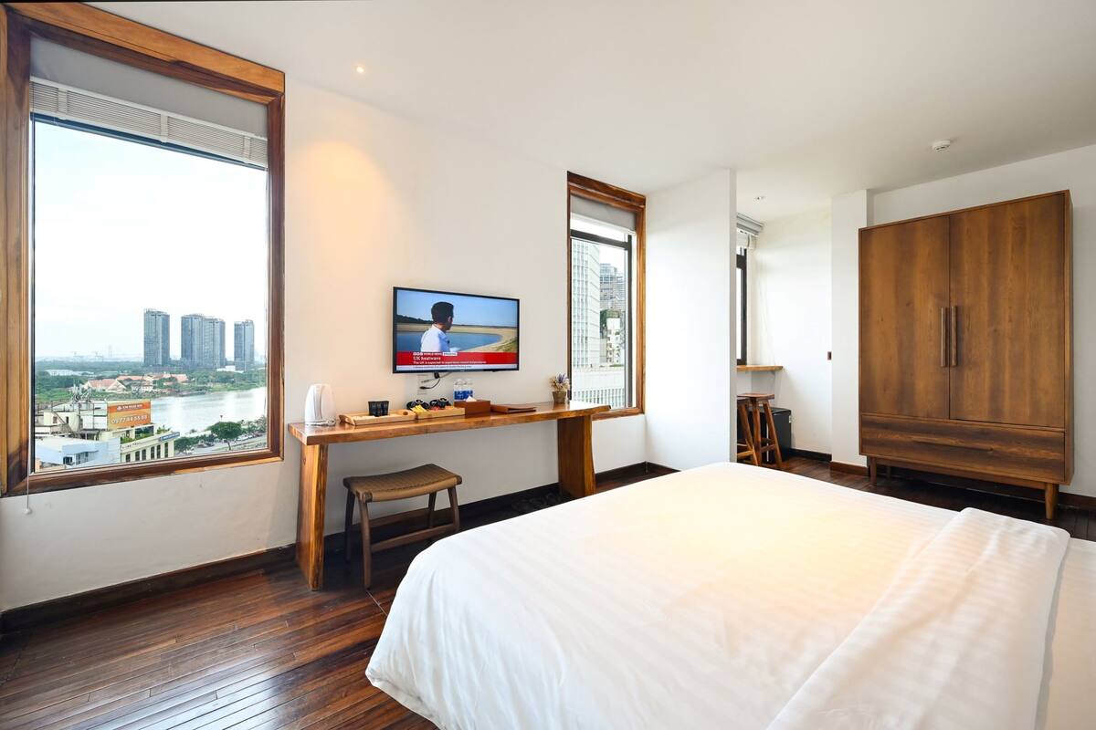 Habitación de hotel con una cama grande, escritorio de madera, televisión y vistas a un paisaje urbano a través de amplios ventanales.