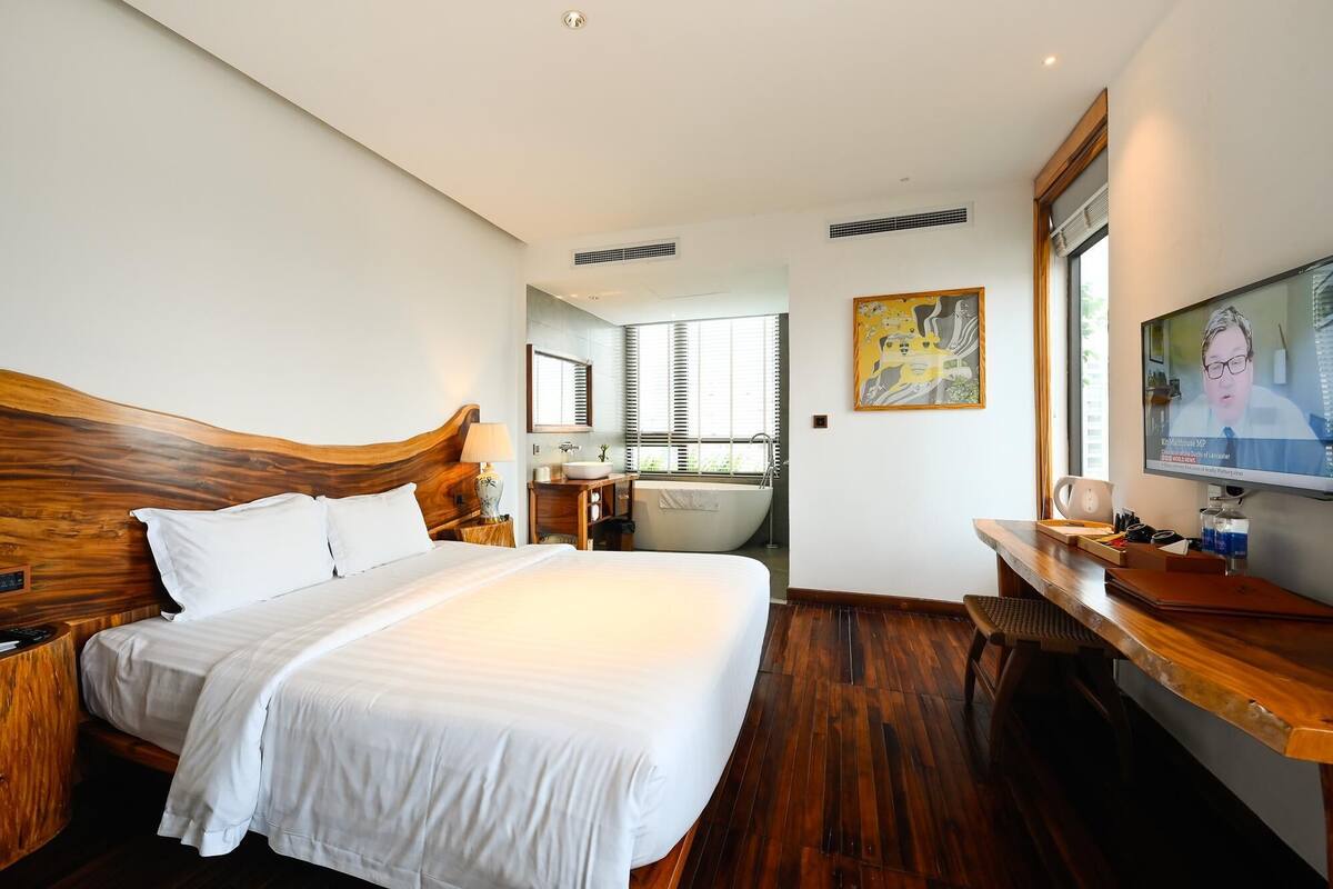 Habitación de hotel con una cama grande, cabecera de madera, tocador con lavabo, bañera y televisor sobre una mesa de madera.
