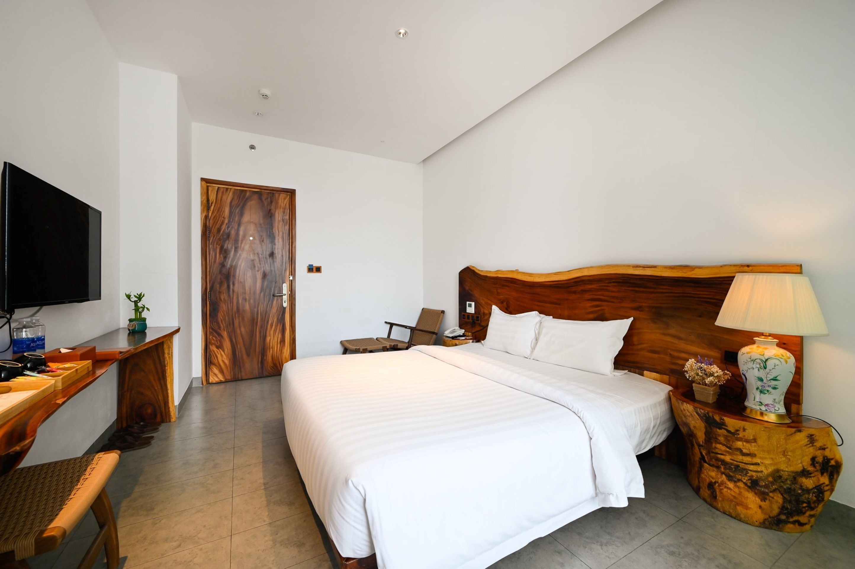 A hotel room with a large bed, wooden headboard, bedside table with a lamp, a TV, and a desk with a chair and a small plant.