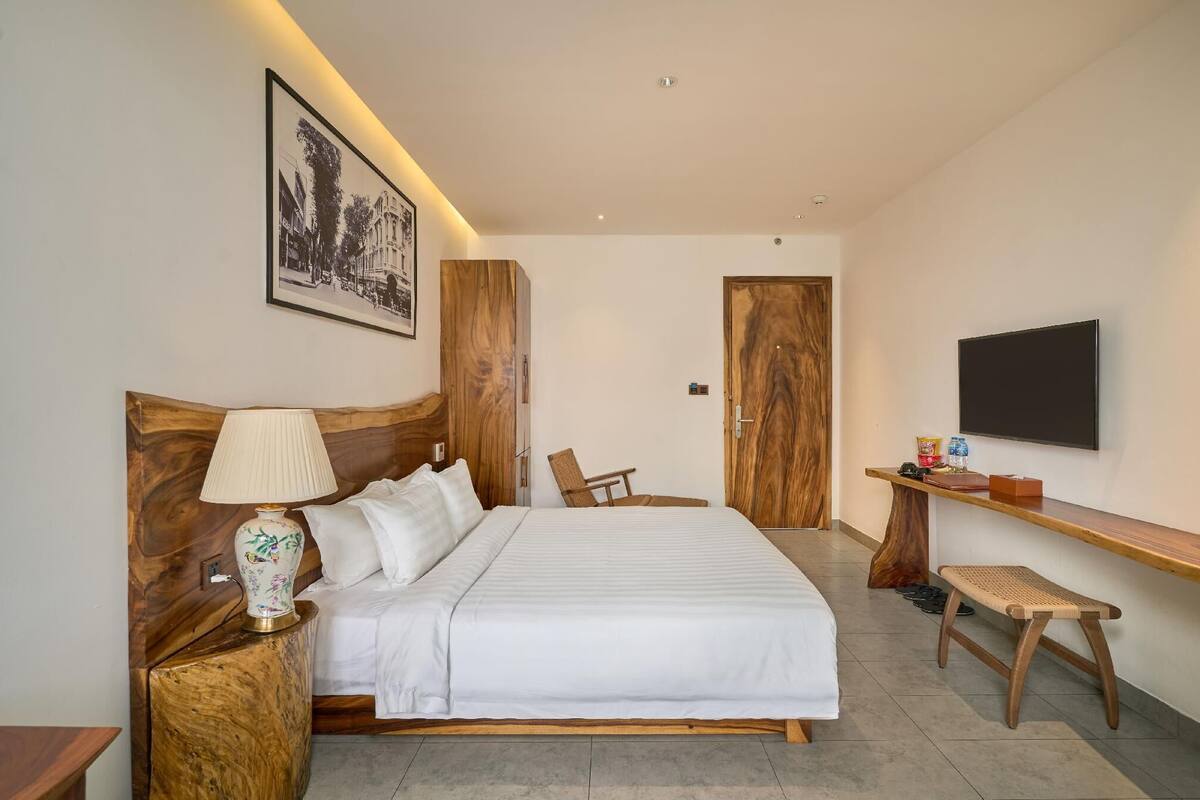 Una habitación de hotel moderna con una cama grande, cabecera de madera, una mesita de noche con lámpara, un televisor montado en la pared y un pequeño banco de madera.