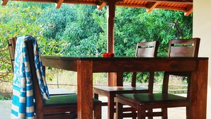 Une table et des chaises en bois, placées à l’extérieur, sous un toit en tôle ondulée. Une des chaises, de couleur verte, est recouverte d’un tissu à motifs bleus et blancs.