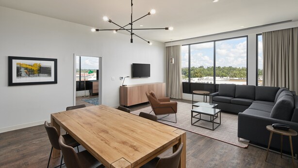 A modern living room with a wooden dining table, a black sofa, and a large window offering a city view. - Hotel Indigo Tallahassee - Collegetown by IHG (Tallahassee)