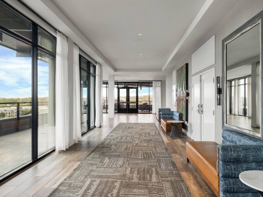 A modern hotel corridor with large windows, a patterned carpet, and seating areas.
