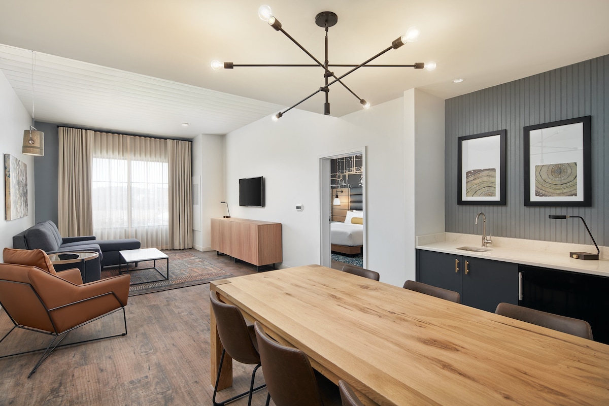 Een moderne hotelkamer met een houten eettafel, bruine leren stoelen, een bank en een kitchenette.