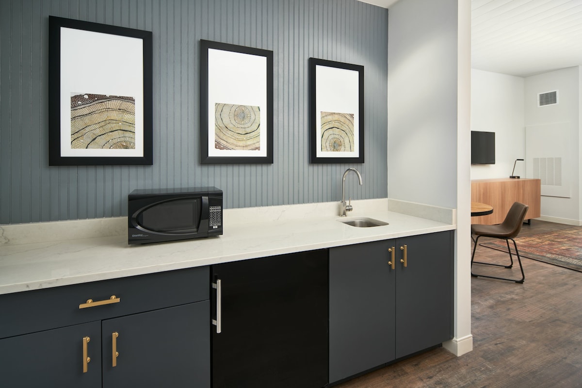 Een moderne keuken met een magnetron, spoelbak en kasten. Drie kunstwerken hangen in lijstjes aan de muur.