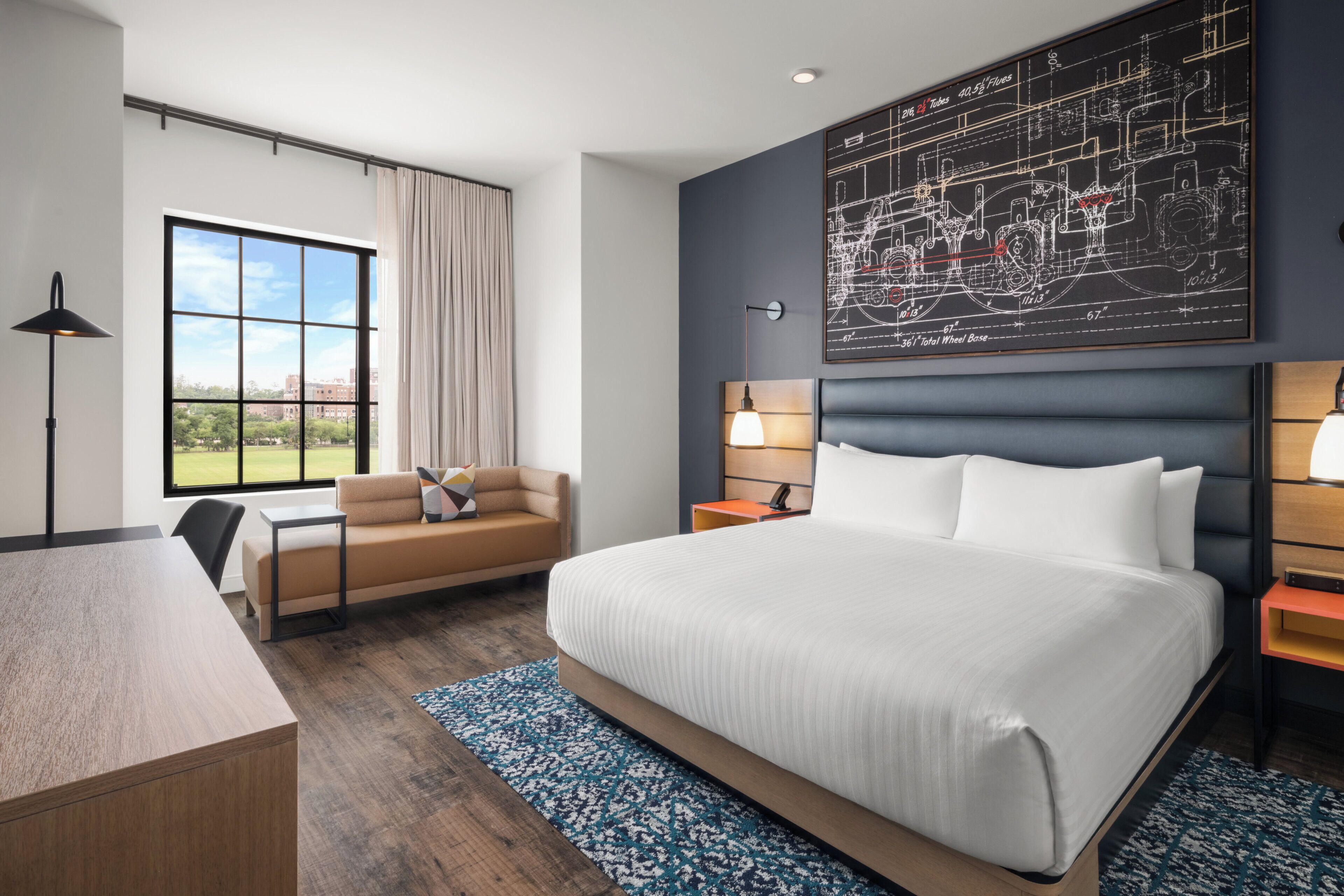 Quarto de hotel moderno com uma cama grande, uma escrivaninha, uma cadeira, um sofá, uma janela com vista e uma obra de arte na parede.