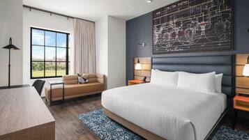 A modern hotel room with a large bed, a desk, a chair, a sofa, a window with a view, and a wall-mounted artwork.