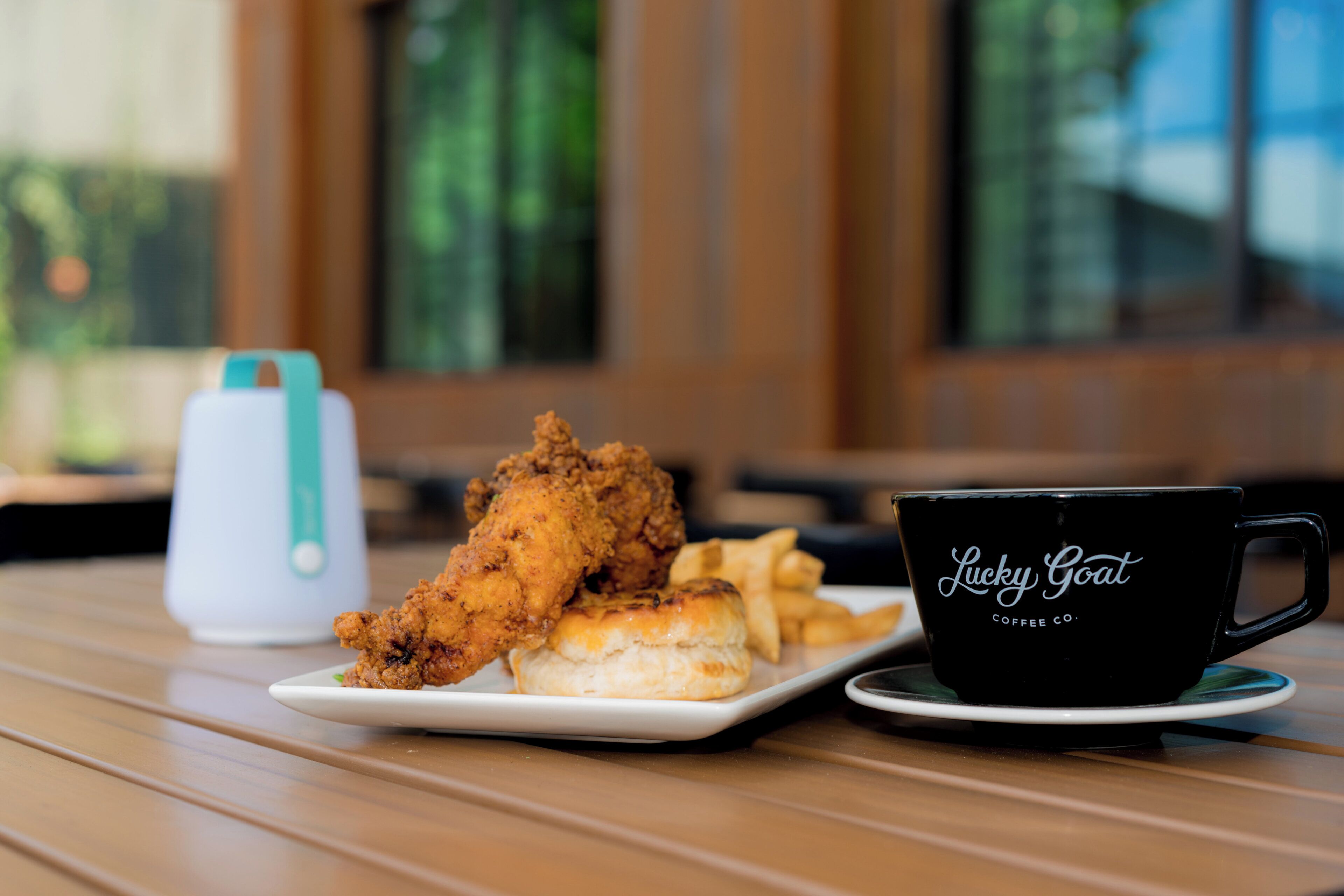Una mesa con un plato de pollo frito, galletas y papas fritas, y una taza de café negro con la inscripción 'Lucky Goat Coffee Co.'.