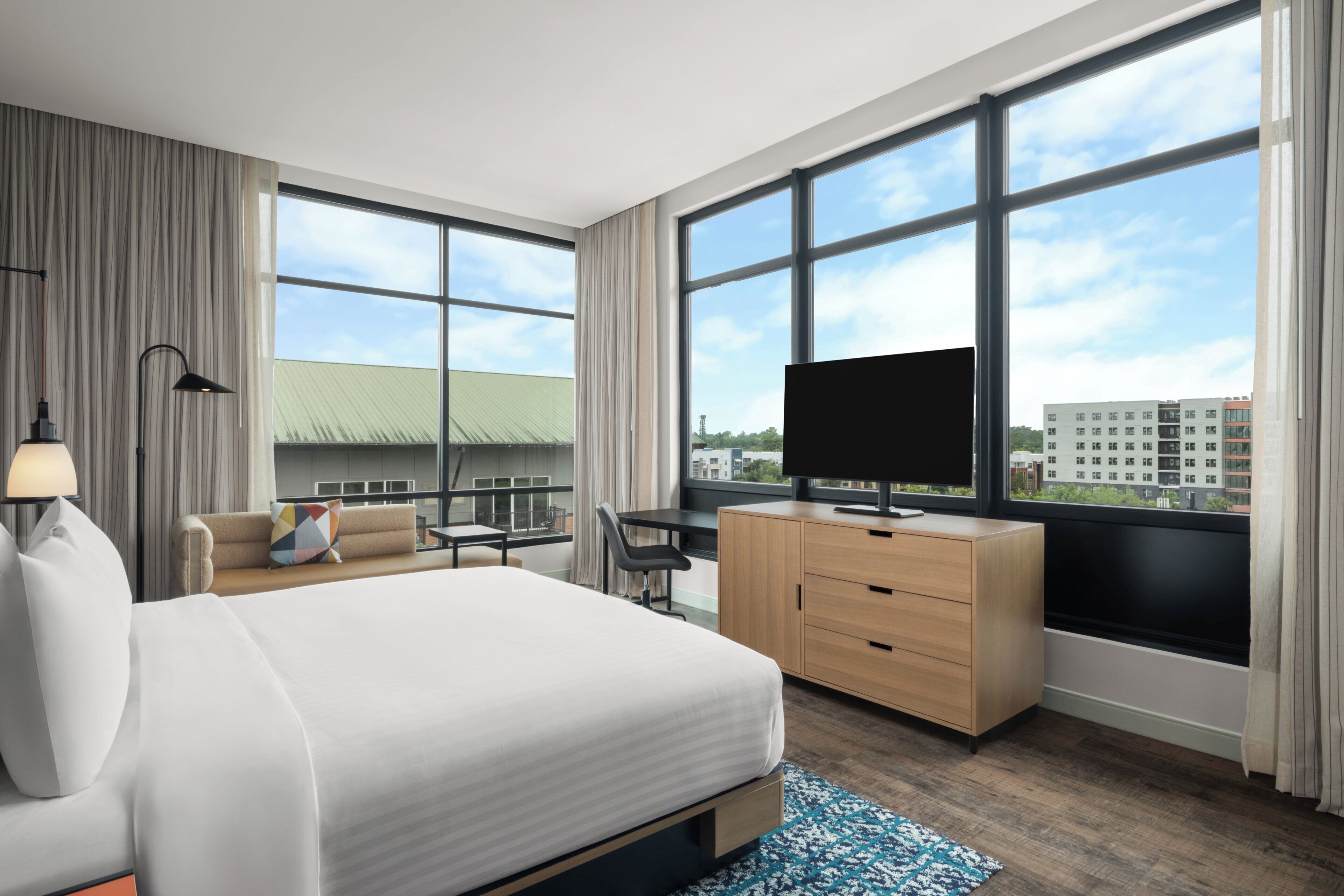 A modern hotel room with a large bed, a desk, a television, and a view of the city through large windows.