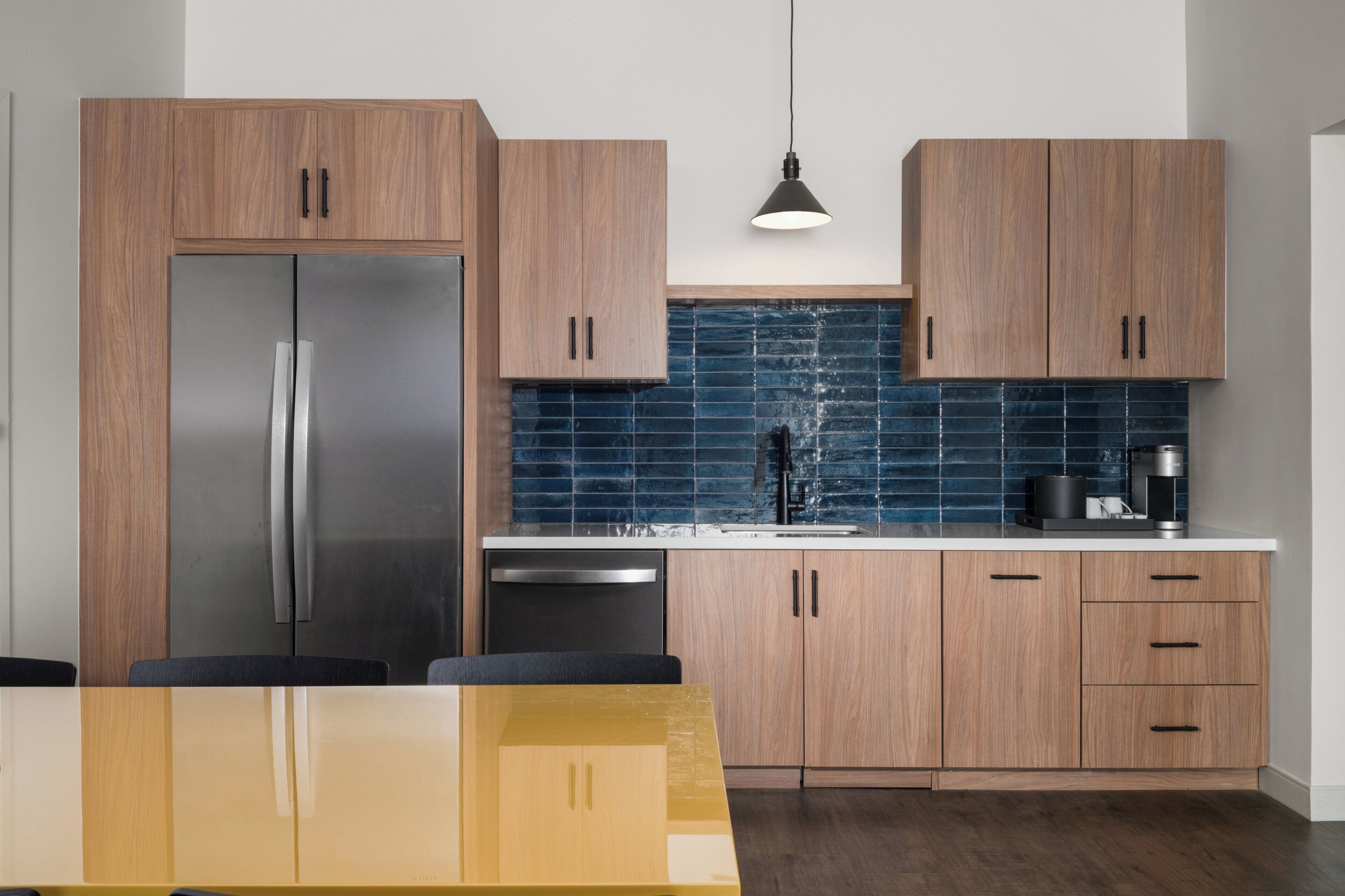 A modern kitchen with wooden cabinets, a stainless steel refrigerator, a blue tiled backsplash, and a hanging pendant light.