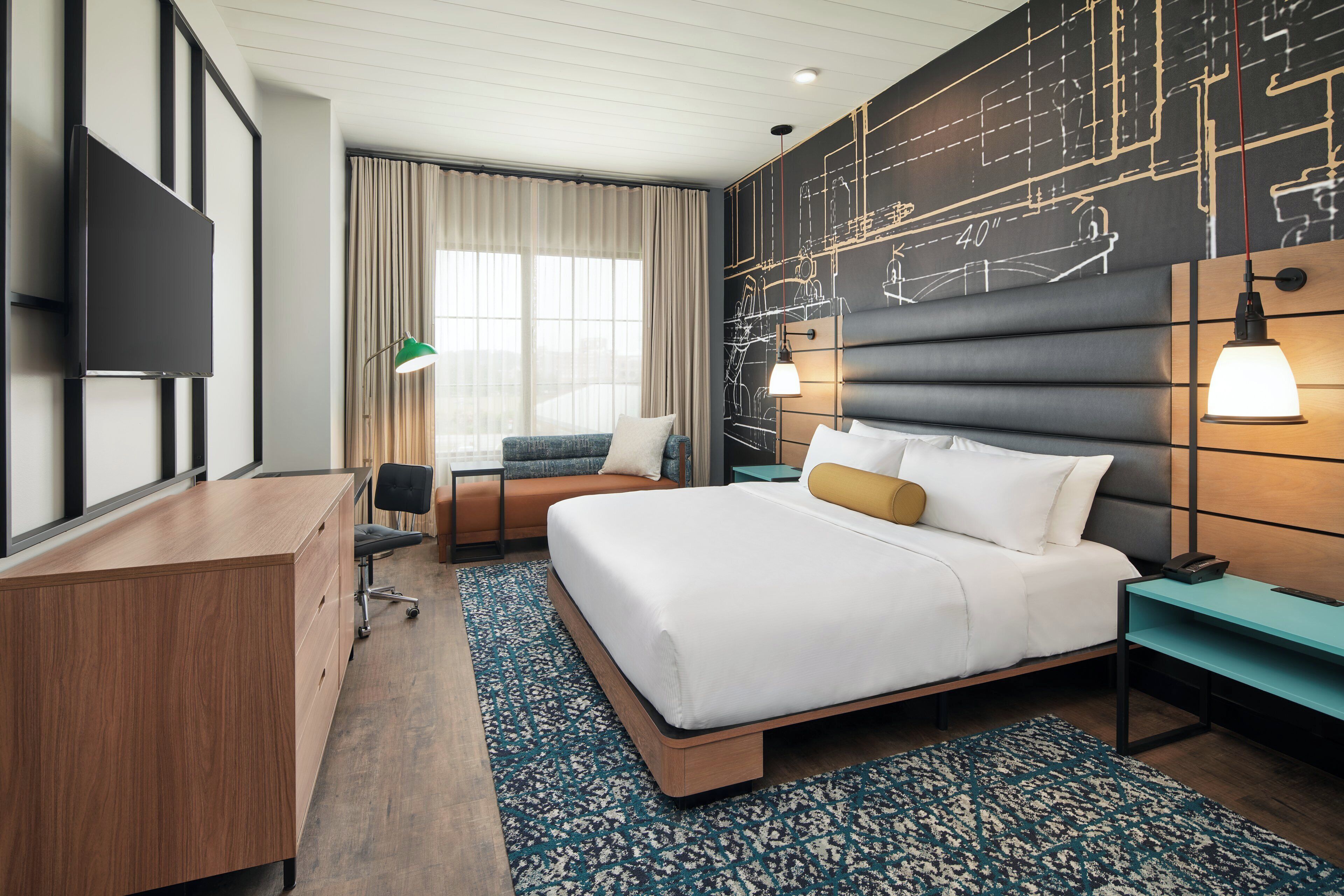 A modern hotel room with a large bed, a desk, a chair, a TV, and a wall with a blueprint design.