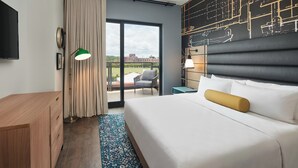 A hotel room with a large bed, a desk, a chair, a balcony with a view, and a wall with architectural drawings.