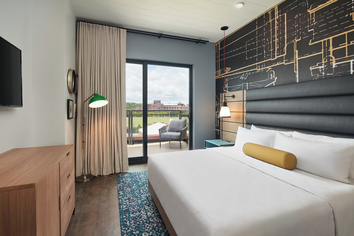 Habitación de hotel con una cama grande, un escritorio, una silla, un balcón con vistas y una pared con dibujos arquitectónicos.