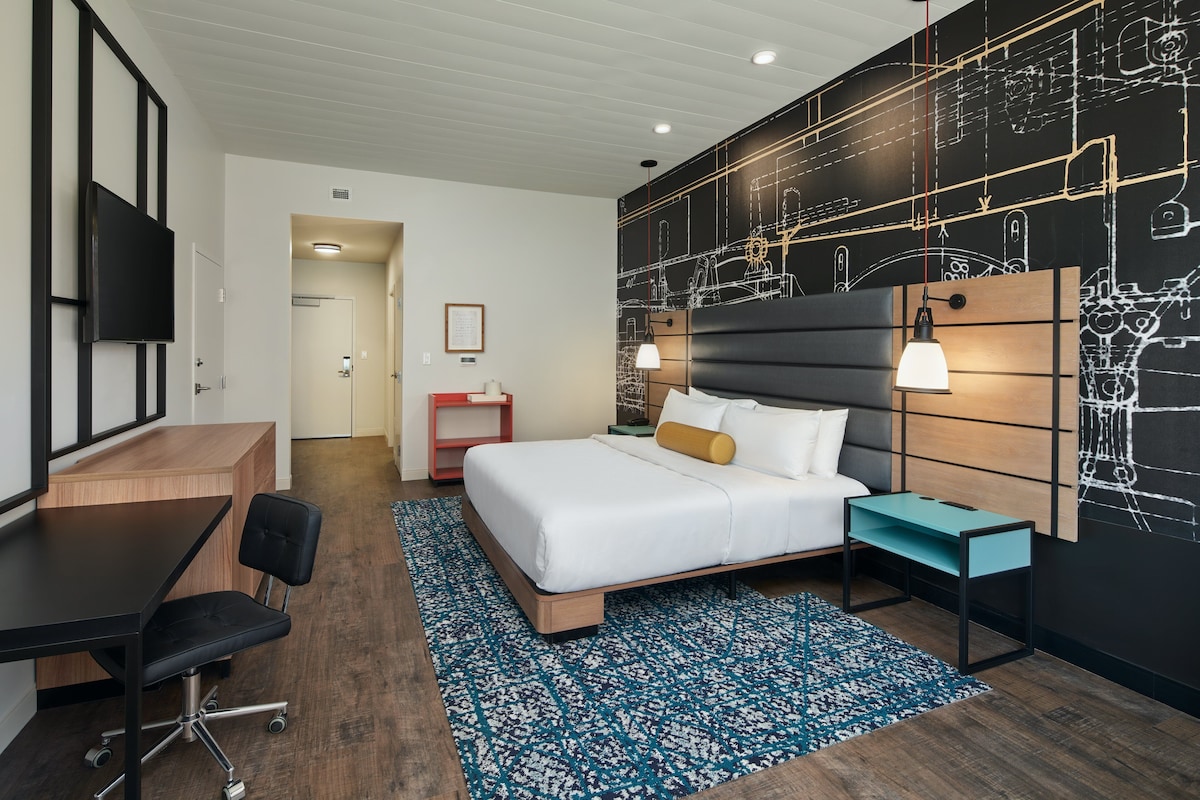 Een moderne hotelkamer met een bed, bureau en stoel, een blauw kamerkleed en een wand met een zwart-wit ontwerp.