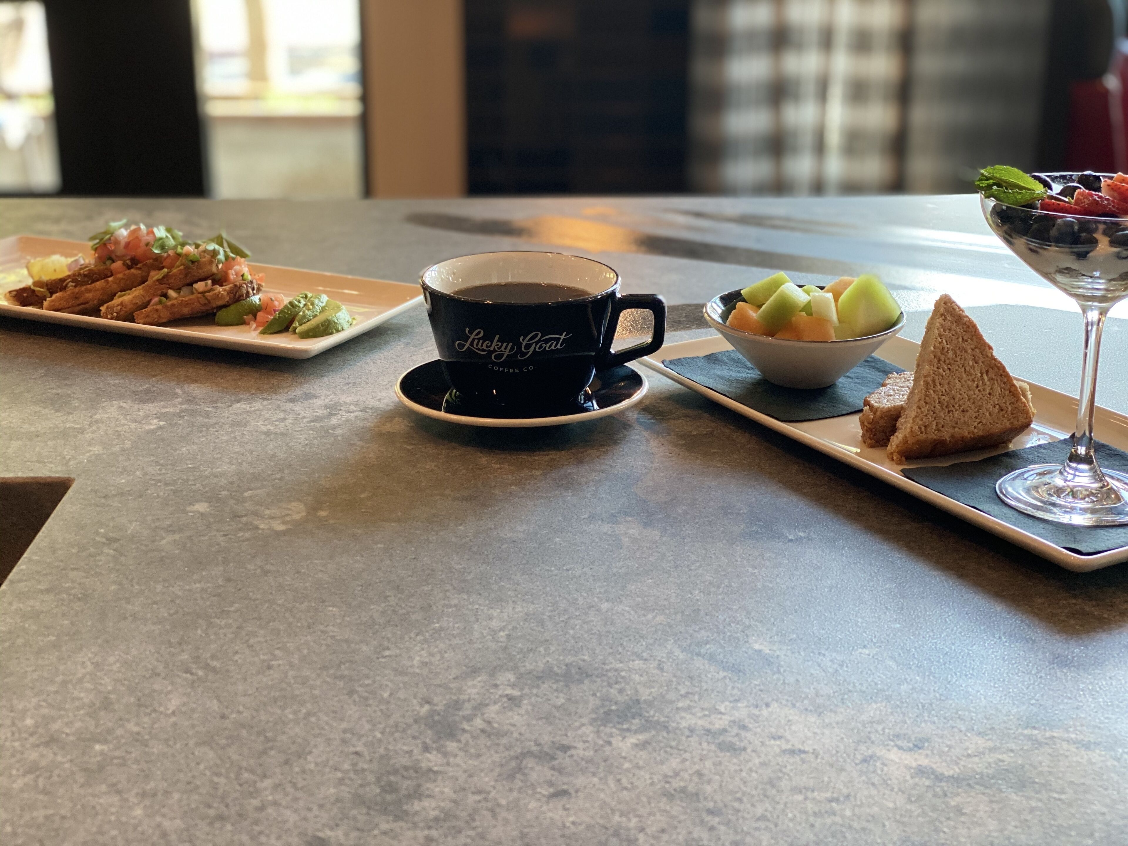 Una mesa con una taza de café, un plato de comida, un cuenco de fruta y un vaso de agua.