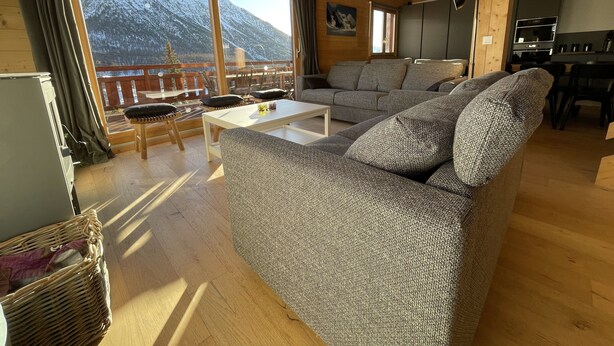 A spacious living room with a large sofa, a coffee table, and a view of snow-capped mountains through large windows. - New charming chalet, 15 to 16 people with panoramic terrace facing south (Montgenèvre)
