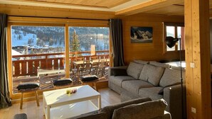 A cozy cabin interior with a wooden ceiling, a sofa, a dining table, and a balcony with a view of a snowy landscape. - New charming chalet, 15 to 16 people with panoramic terrace facing south (Montgenèvre)