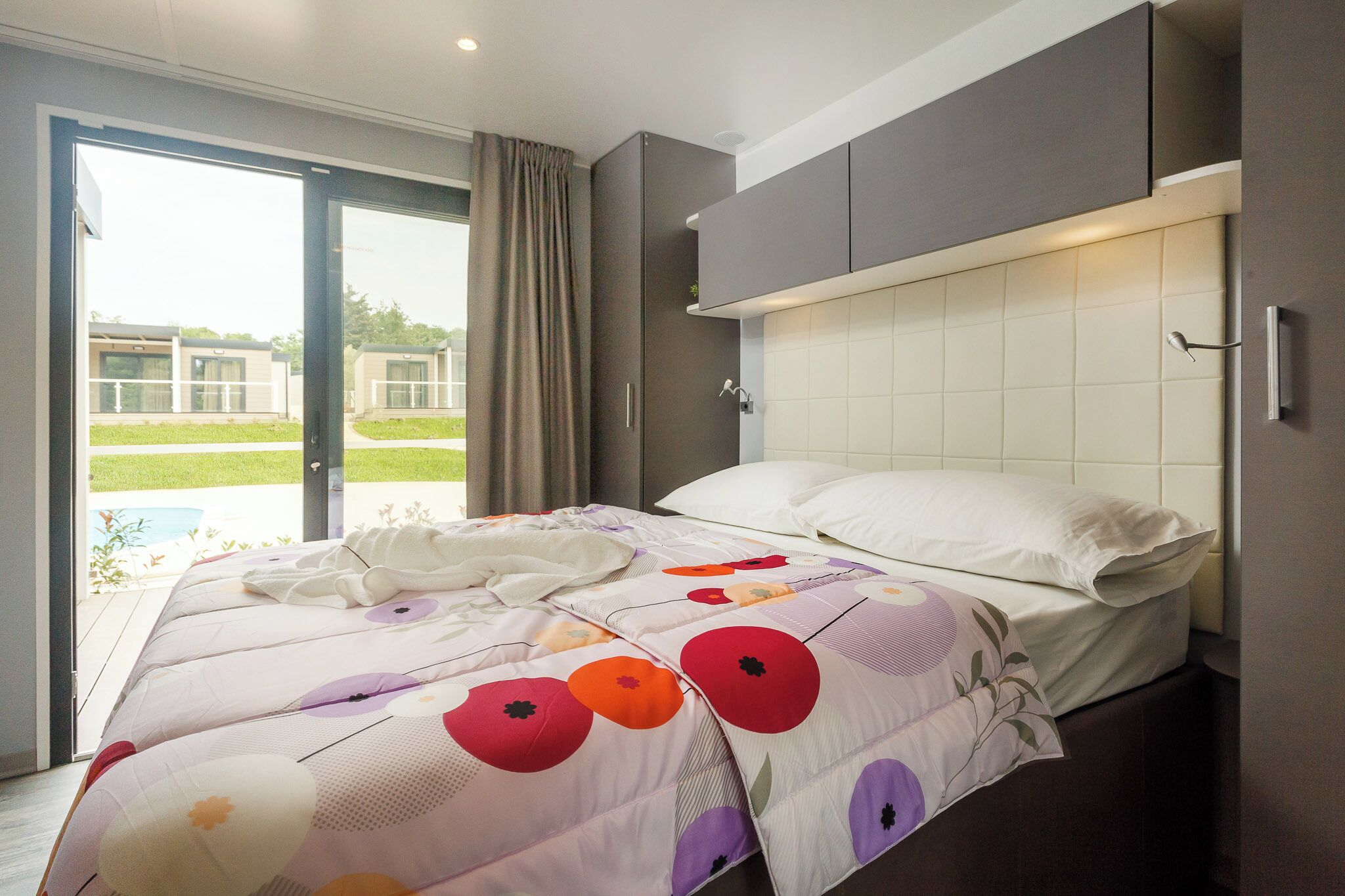 A modern bedroom with a bed, a sliding glass door to a balcony, and a view of a pool.