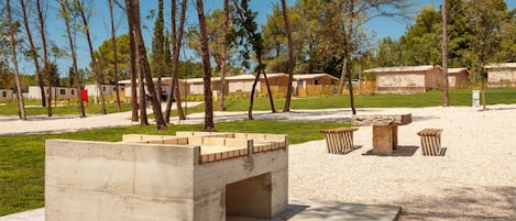 A concrete structure with benches and a stone table in a park-like setting with trees and a row of mobile homes in the background.