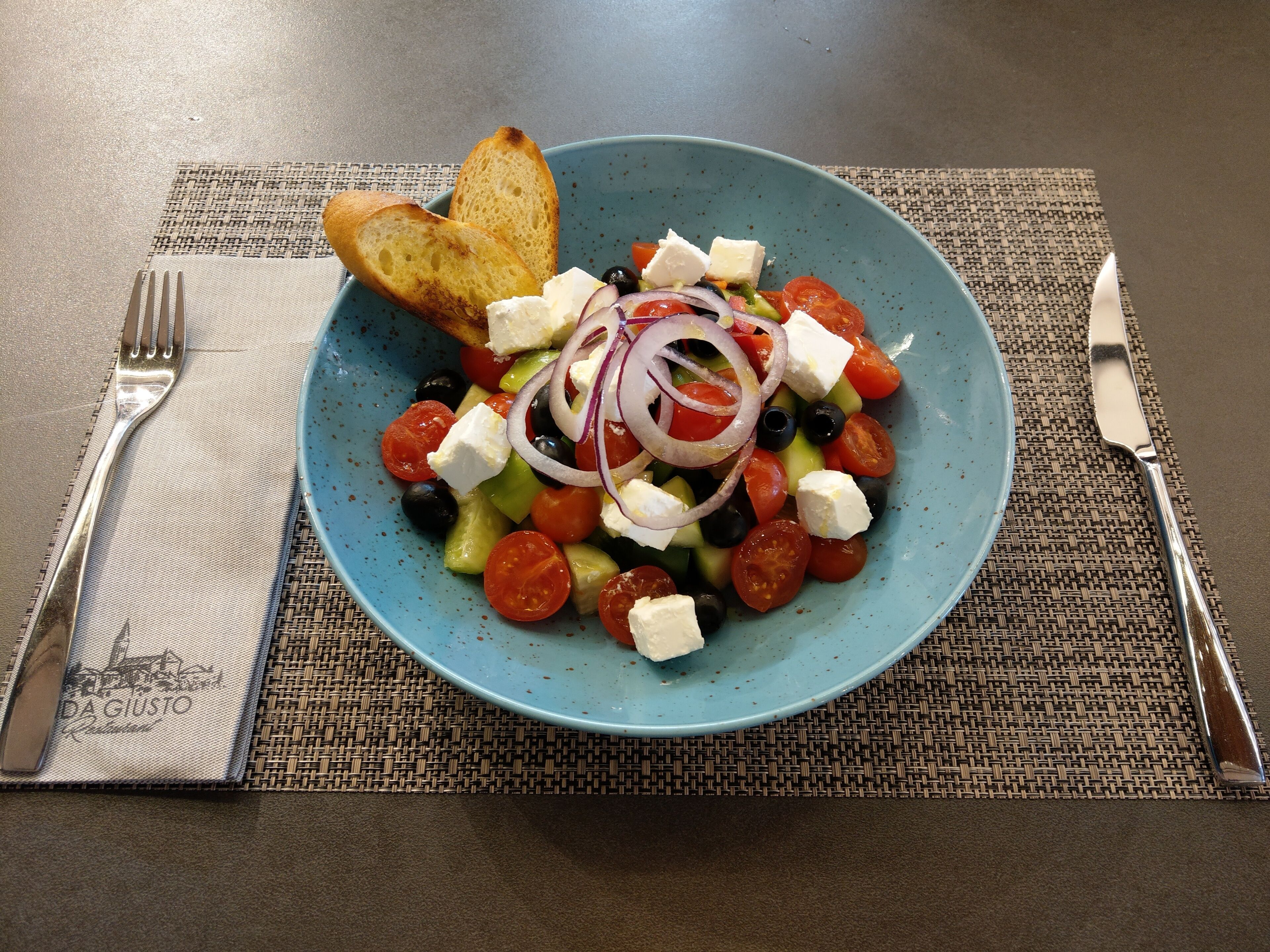 Een blauw bord met een salade, met tomaten, komkommers, olijven en feta, vergezeld van geroosterd brood en een vork en mes op een placemat.