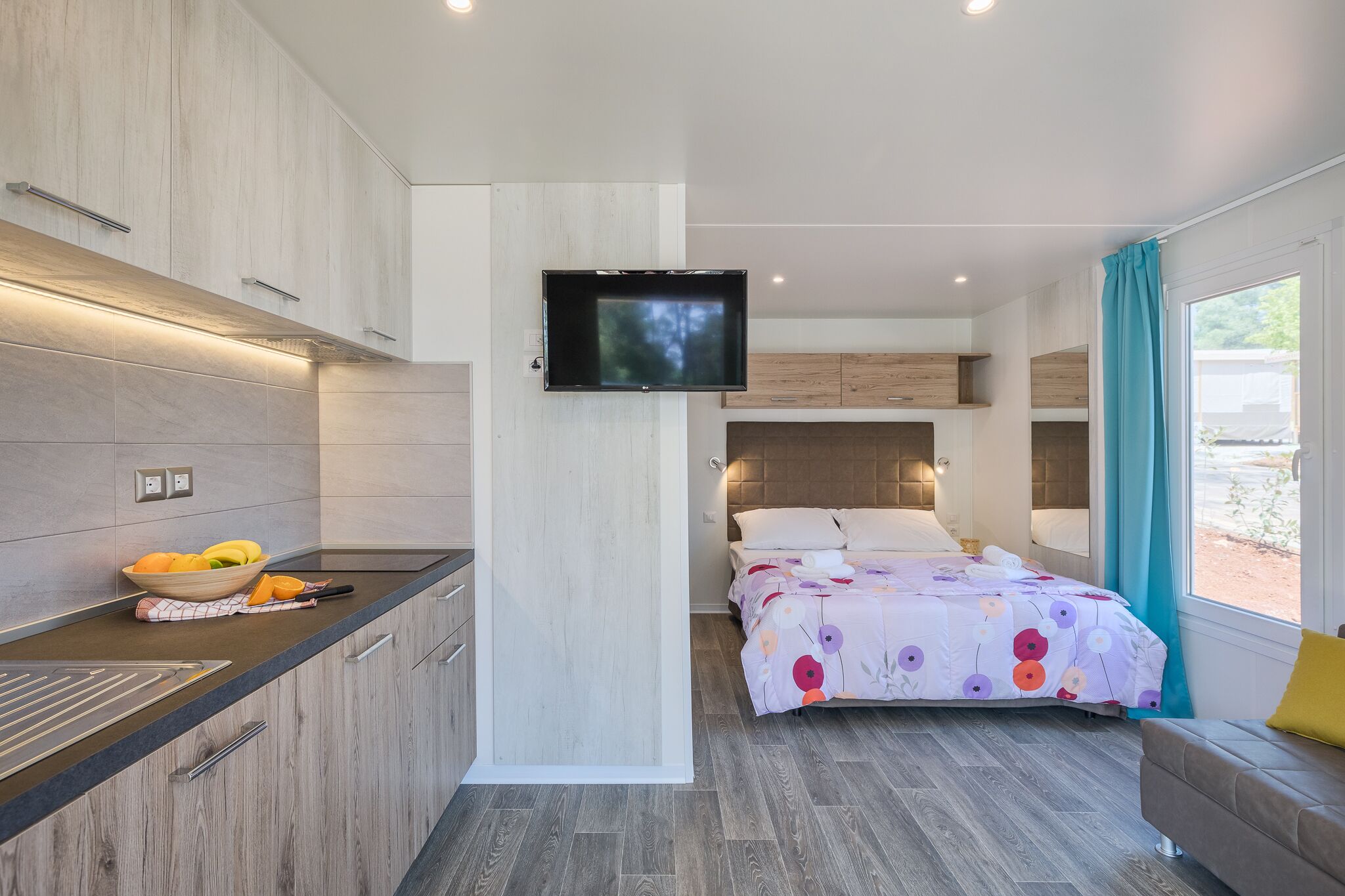 Habitació moderna amb zona de cuina, un llit amb edredó floral i un televisor fixat a la paret.