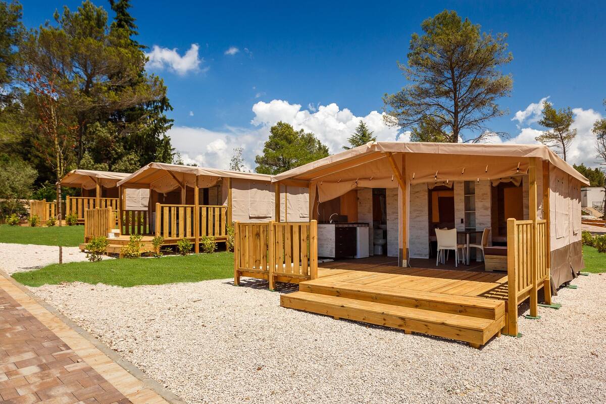 Una hilera de cabañas de madera con asientos exteriores y zona de cocina, ubicadas en un área con césped y árboles al fondo.