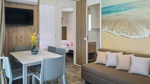 Una habitación de hotel moderna con una televisión de pantalla plana, una mesa de comedor con sillas, un sofá con cojines y un mural de playa en la pared.