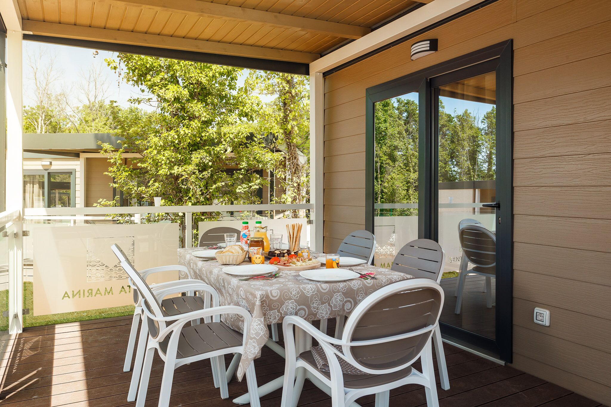 An outdoor patio with a table set for dining, surrounded by white chairs, and a glass door leading to a covered area.