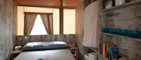 A compact bedroom with a bed, a shelf with books, and a window with curtains.