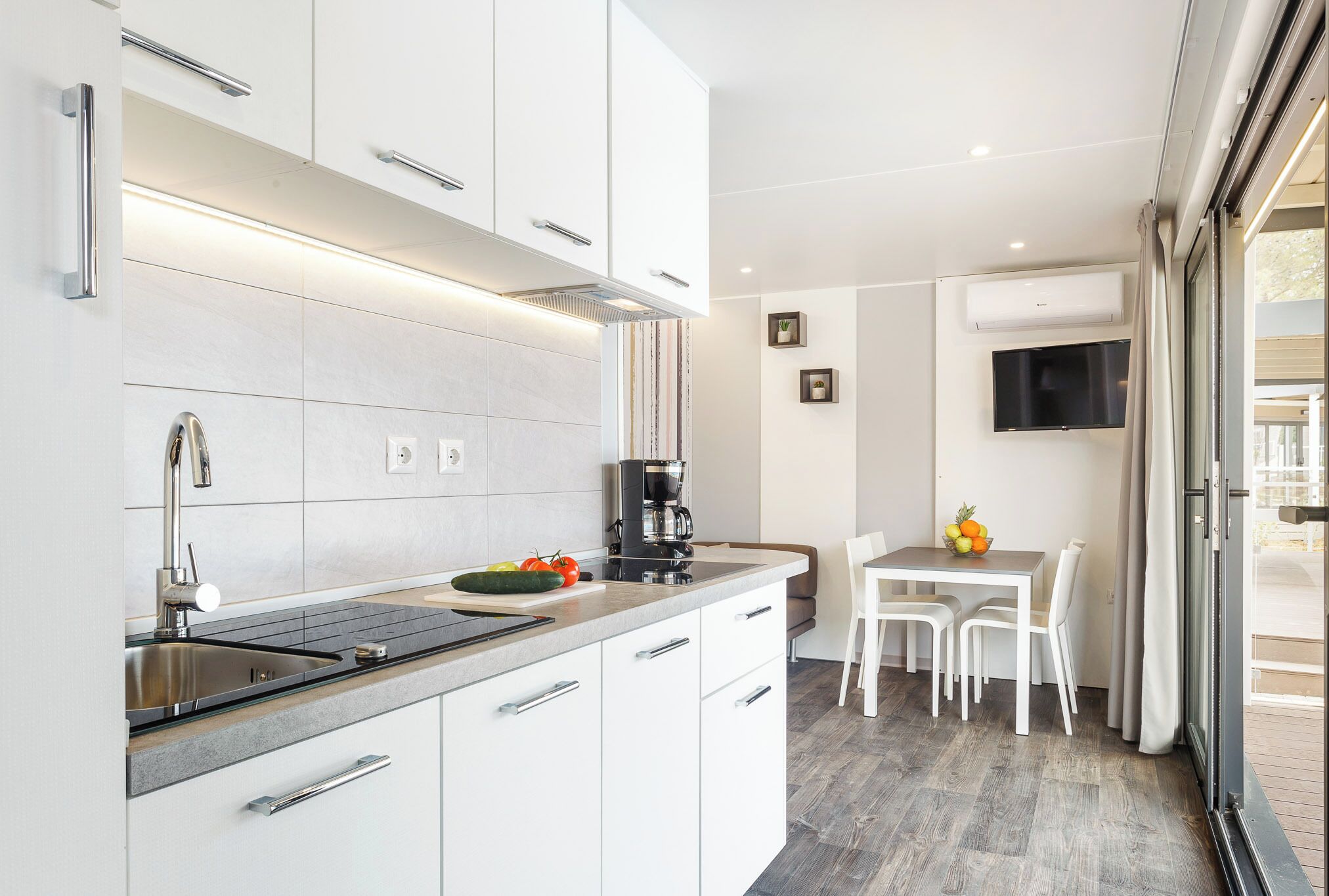 Una cocina moderna con armarios blancos, un lavamanos de acero inoxidable y una cafetera. Hay un comedor con mesa y sillas, y un televisor montado en la pared.