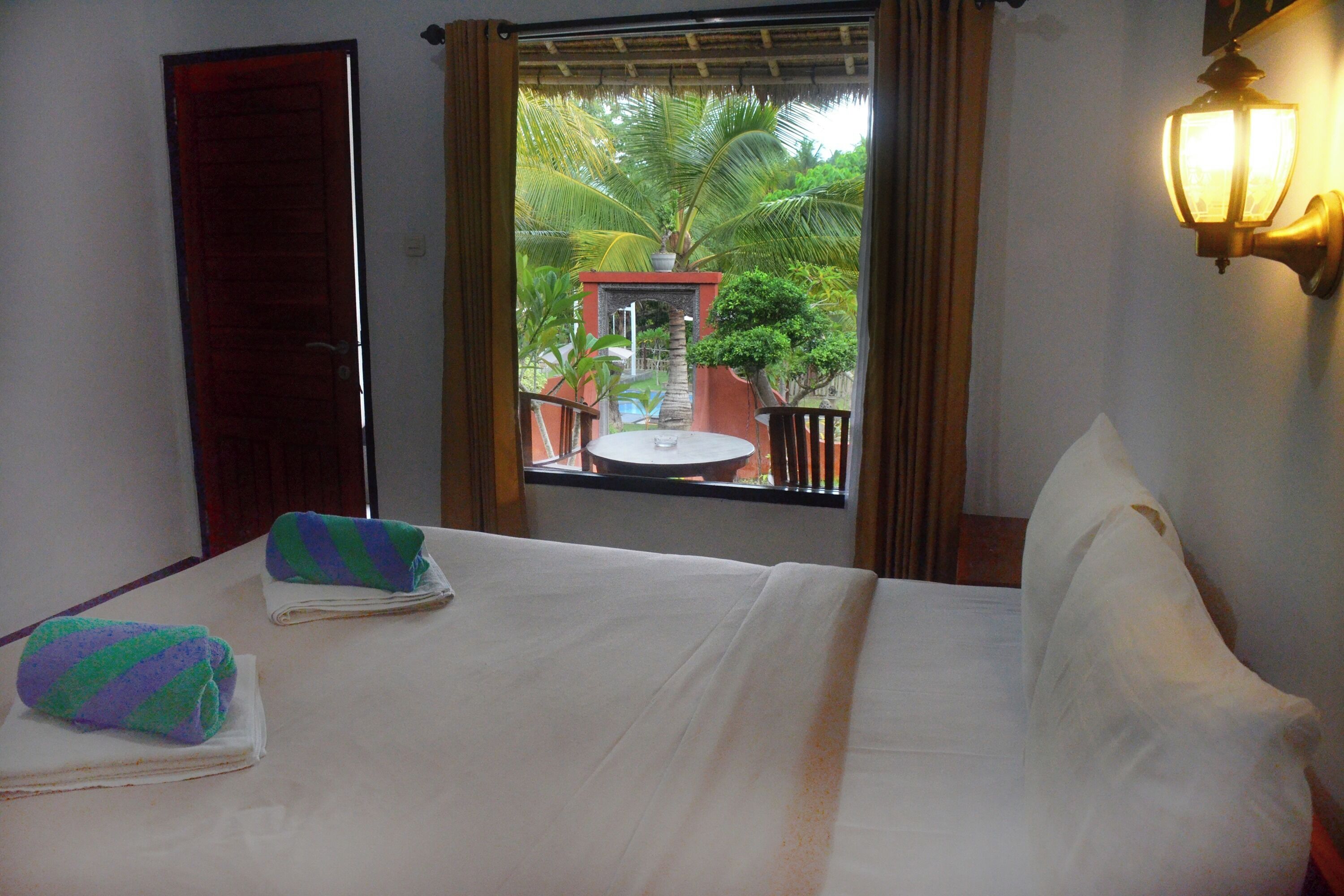 A hotel room with a bed, a window with a view of a palm tree and a pool, and a wall-mounted light fixture.