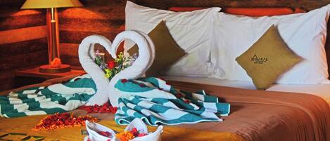 A neatly made bed with a heart-shaped towel arrangement, a lamp, and a wooden headboard.