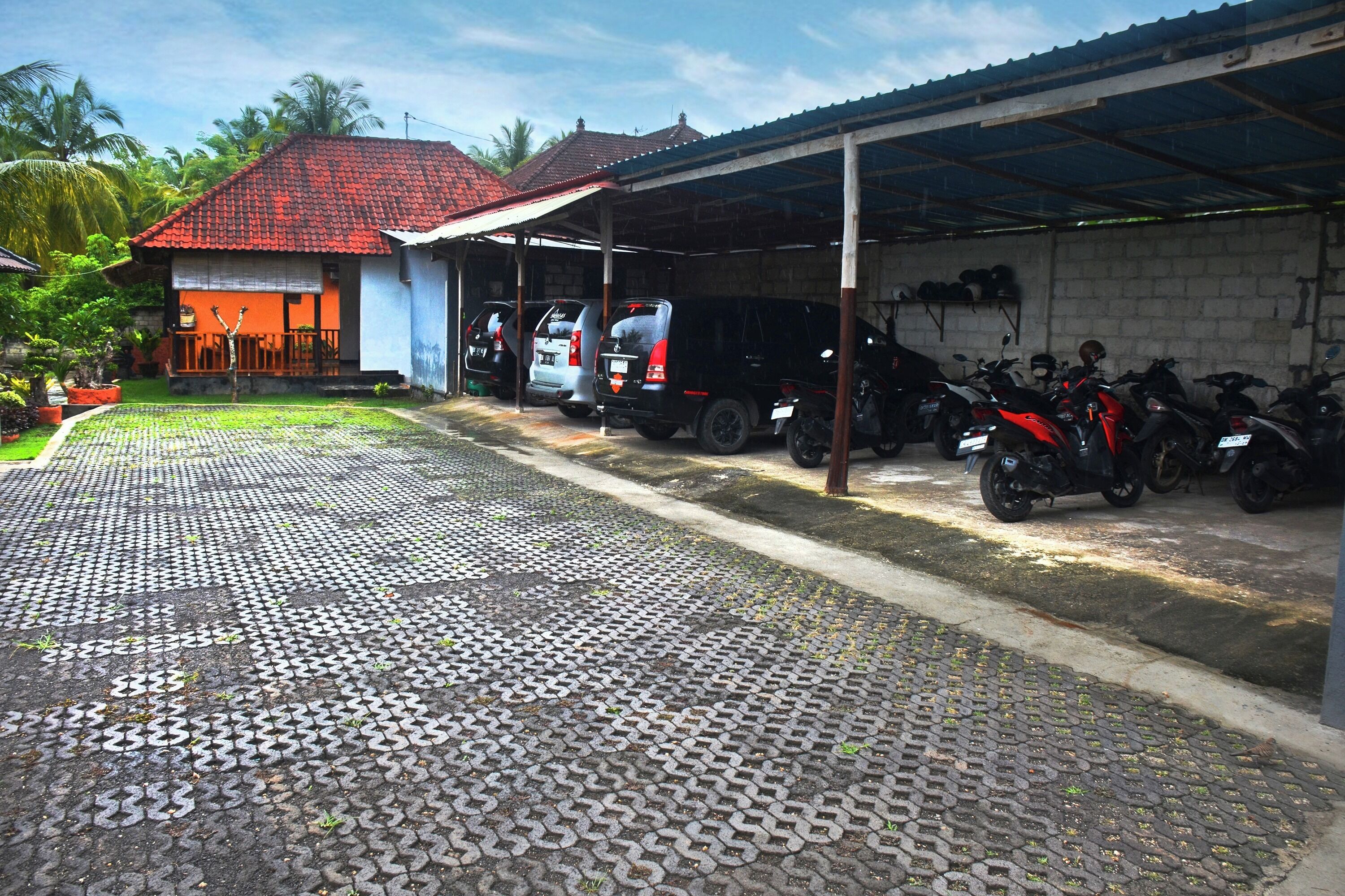 Un'area di parcheggio coperta con pavimento piastrellato, un edificio con tetto rosso e diversi veicoli parcheggiati, tra cui automobili e motocicli.