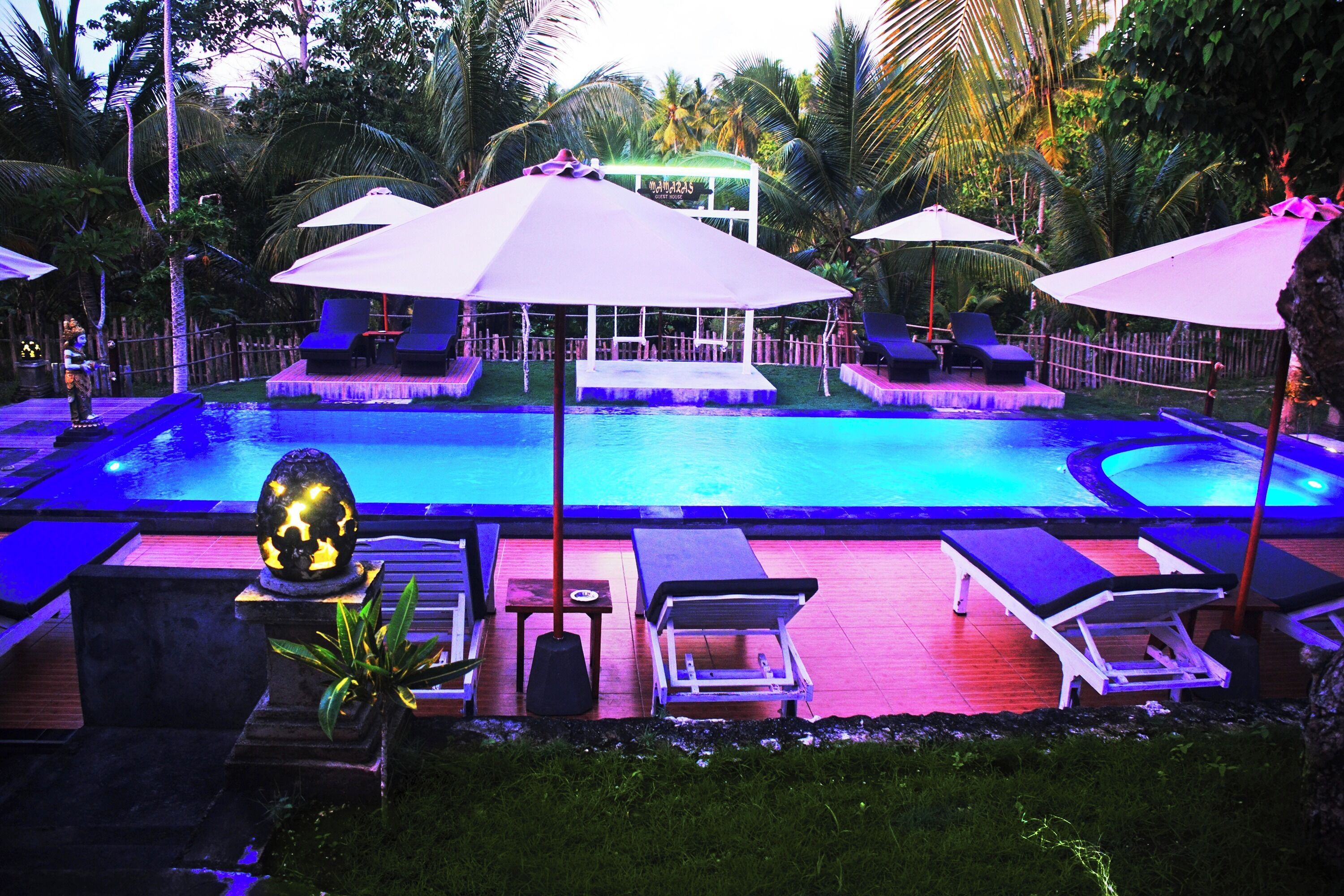 Un área de piscina con sillones de descanso y sombrillas, rodeada de palmeras.