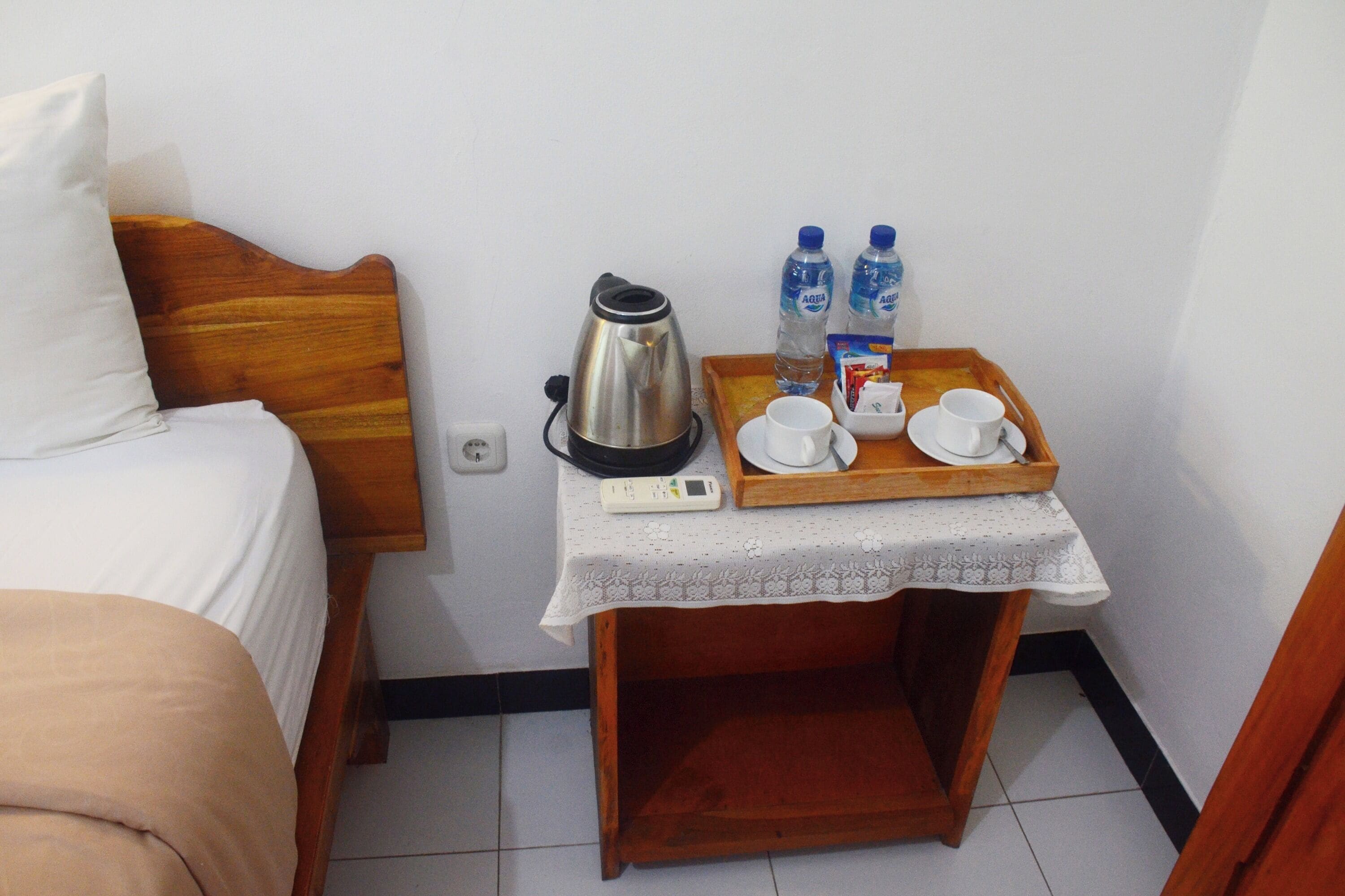 Cama bem arrumada com cabeceira de madeira, mesa de cabeceira com chaleira, duas garrafas de água, bandeja com xícaras e uma caixa, e uma tomada na parede.
